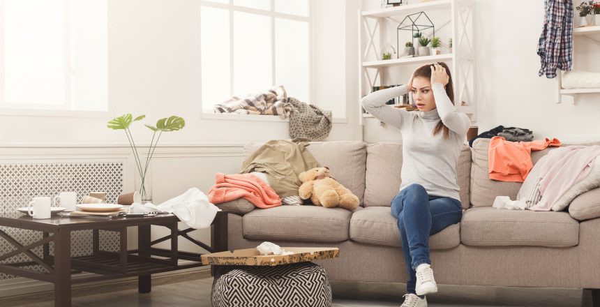 Desperate helpless woman sitting on sofa in messy living room. Young girl surrounded by many stack of clothes. Disorder and mess at home, copy space